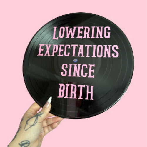 Vinyl record with text 'Lowering Expectations Since Birth' held by a hand on a pink background